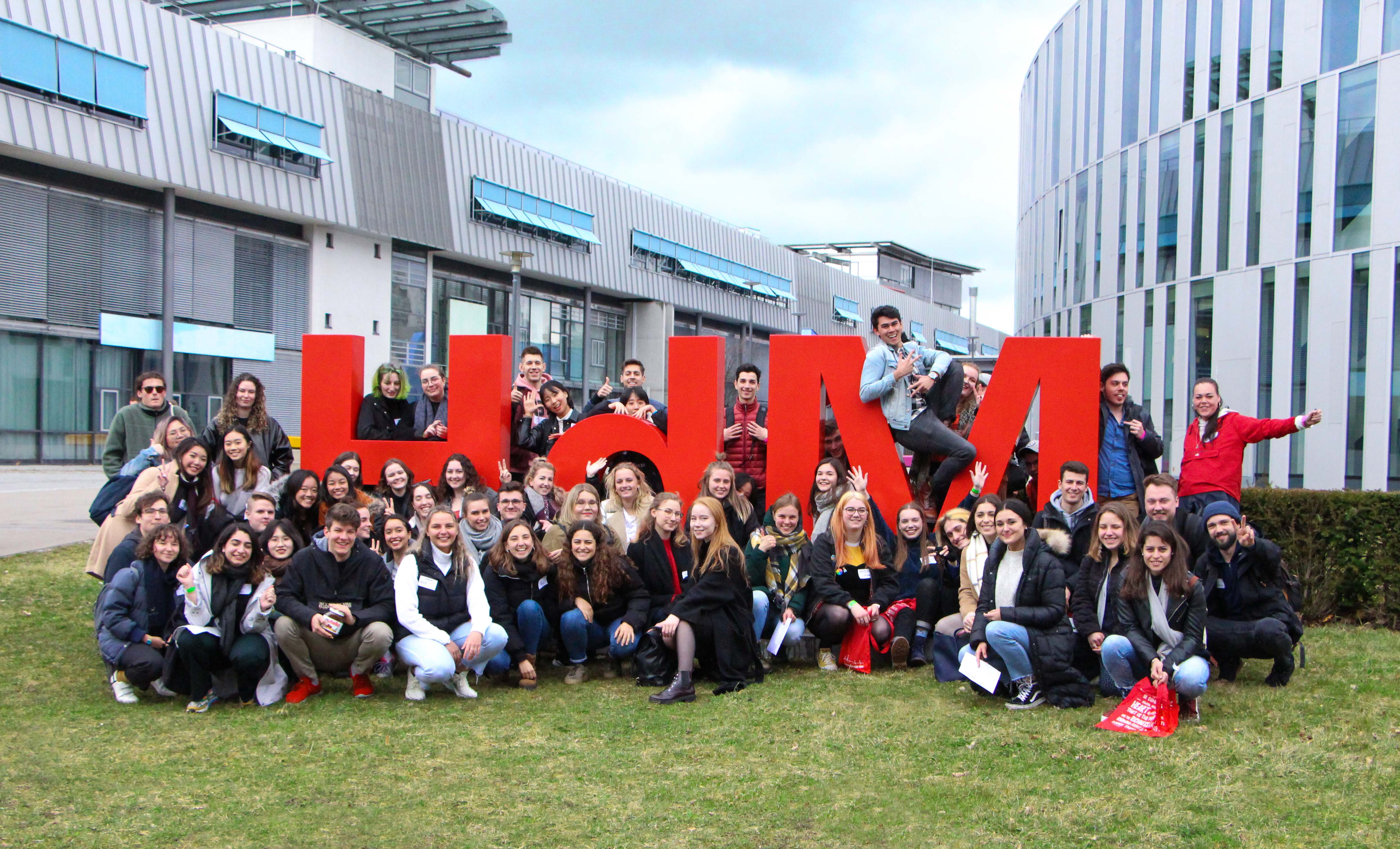 International students standing in front of the HdM logo looking happy